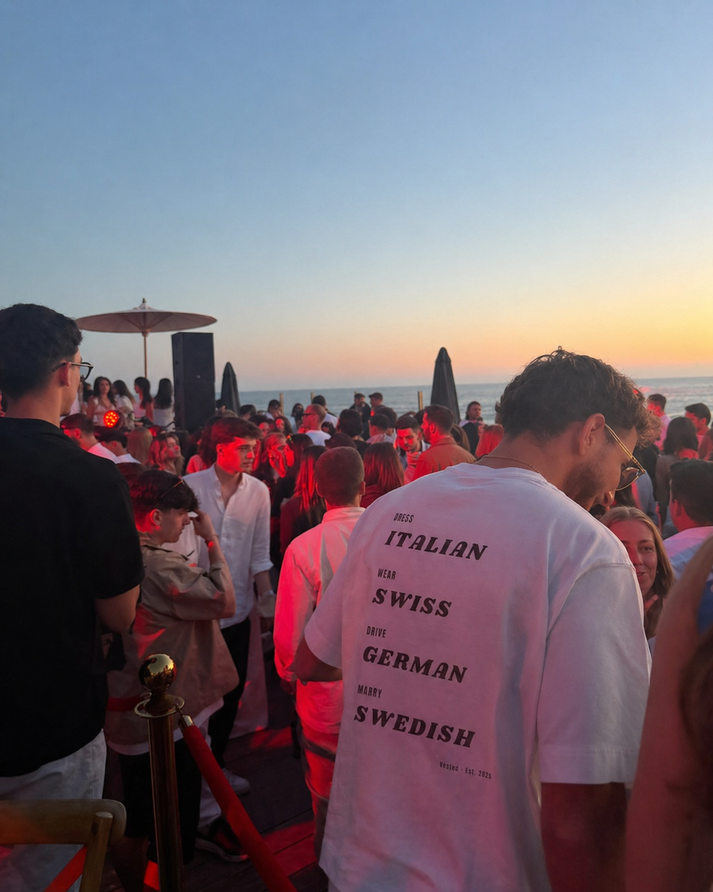 At sunset by the ocean, a crowd in white gathers outdoors. In front, a man wears the DRESS, WEAR, DRIVE, MARRY T-Shirt—a heavyweight cotton tee with bold statement text listing several European nationalities.