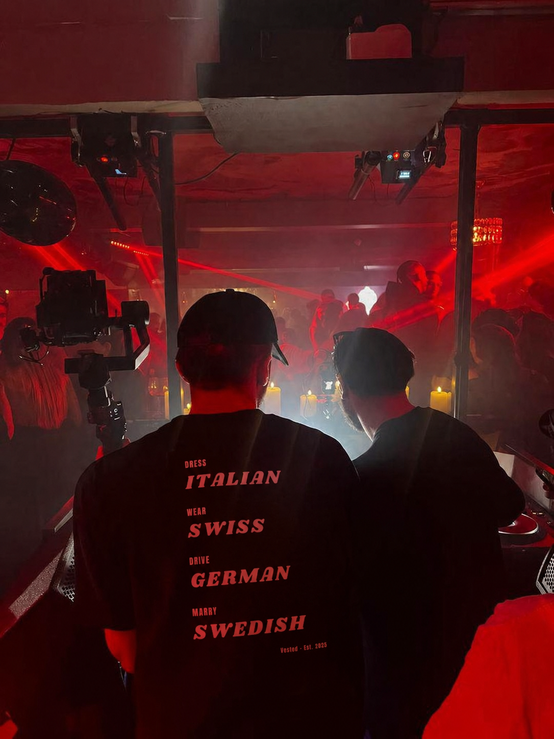 Two DJs in black caps and shirts energize the crowd at a red-lit nightclub. One wears the "DRESS, WEAR, DRIVE, MARRY" T-Shirt, featuring bold statement typography.